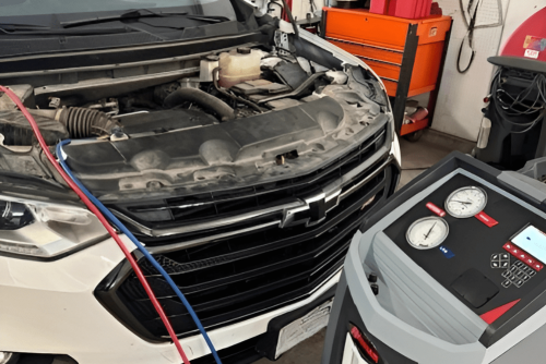 Auto A/C repair & heating service in progress on a white Chevrolet with the hood open, connected to a refrigerant recovery and recharge machine inside a Belton Transmission & Complete Auto Repair in Belton, MO.