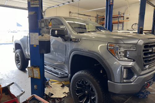Preventative Maintenance in Belton, MO by Belton Transmission & Complete Auto Repair. Image of a silver GMC Sierra on a hydraulic lift inside an auto repair shop, showing a clear view of suspension and lift equipment in use.